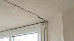 Fissure fine visible dans le mur au-dessus d’une fenêtre d’appartement, photographiée en lumière naturelle.
